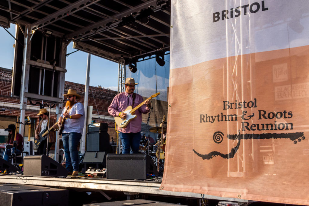 Wide shot of the band 49 Winchester performing on stage with a Bristol Rhythm and Roots Reunion banner in the foreground. Three men hold guitars while wearing cowboy hats while a woman sits at an instrument with a cowboy hat on as well.