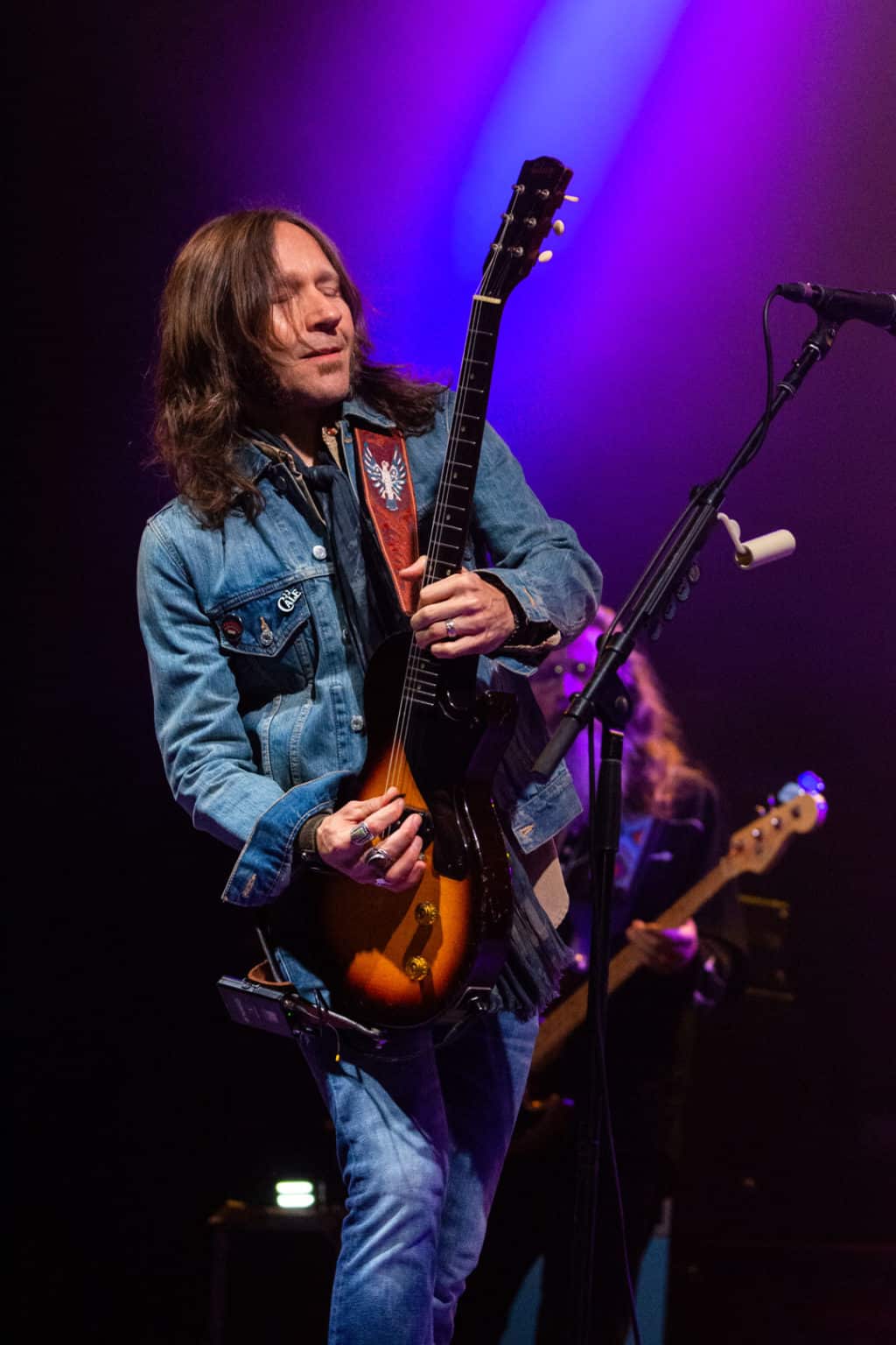 Medium shot of Blackberry Smoke smiling with eyes closed while playing an electric guitar with purple lights shining in the background