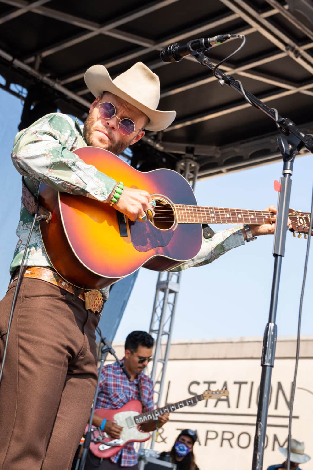 Medium shot of Charlie Crockett holding his acoustic guitar high up as he picks it. He is wearing a cowboy hat, round sunglasses, colorful jewelry, and a belt with white horses on it and a card hand belt buckle.
