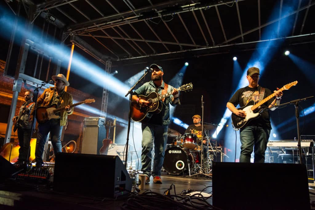 Wide shot of Folk Soul Revival playing on stage at night with beams of blue light shining in multiple directions. Four men can be seen standing and holding guitars with a dummer playing at the back of the stage.