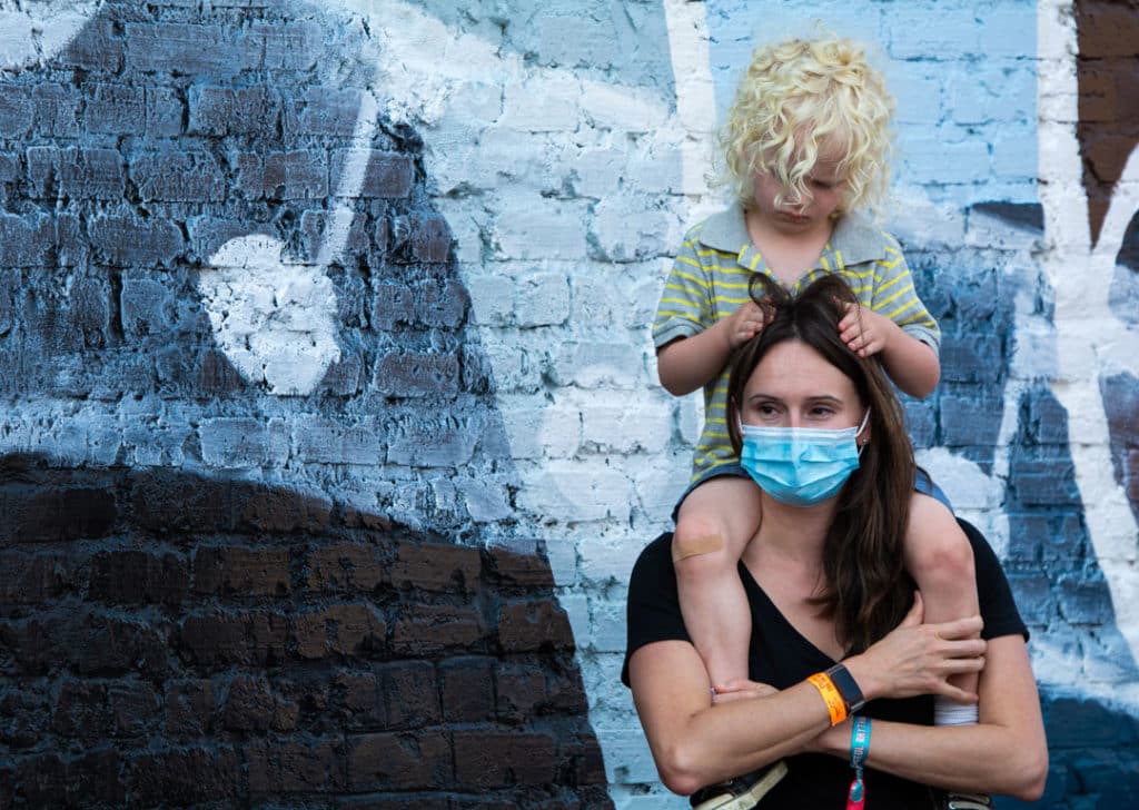A woman smiles under her medical mask while standing in front of a mural while a child sits on her shoulders and pulls at her hair. The woman is wearing a festival pass bracelet.