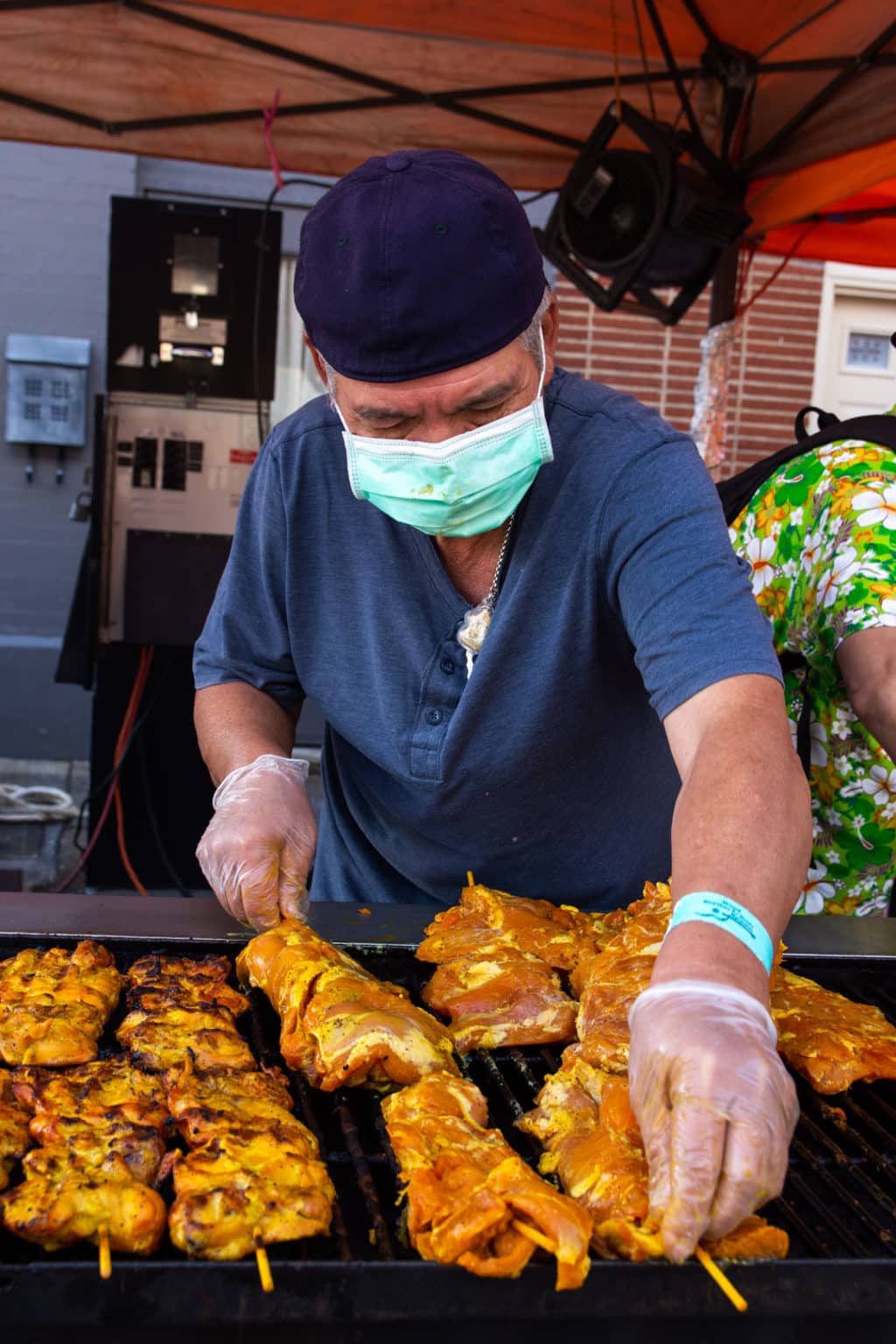 A vendor cooks meat on a grill while wearing a backwards hat and a medical mask.