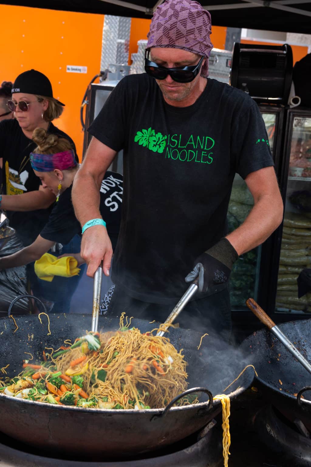 A vendor cooking noodles and vegetables in a large wok while wearing goggles, a purple bandana on their head, and a shirt that says "Island Noodles."