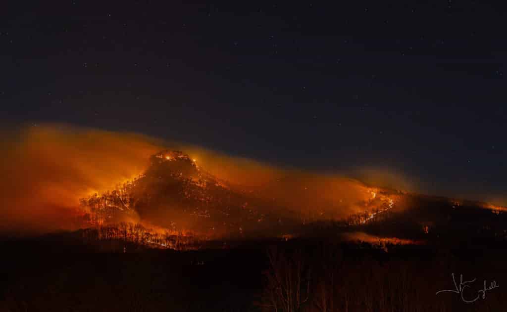View of Pilot Mountain at night covered in bright orange flames as smoke blows off the mountain