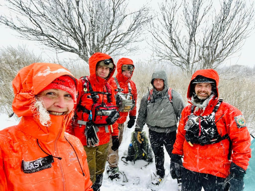 Rescuers from Haywood County Search and Rescue stand and smile in the snow storm while wearing bright orange gear and safety equipment.
