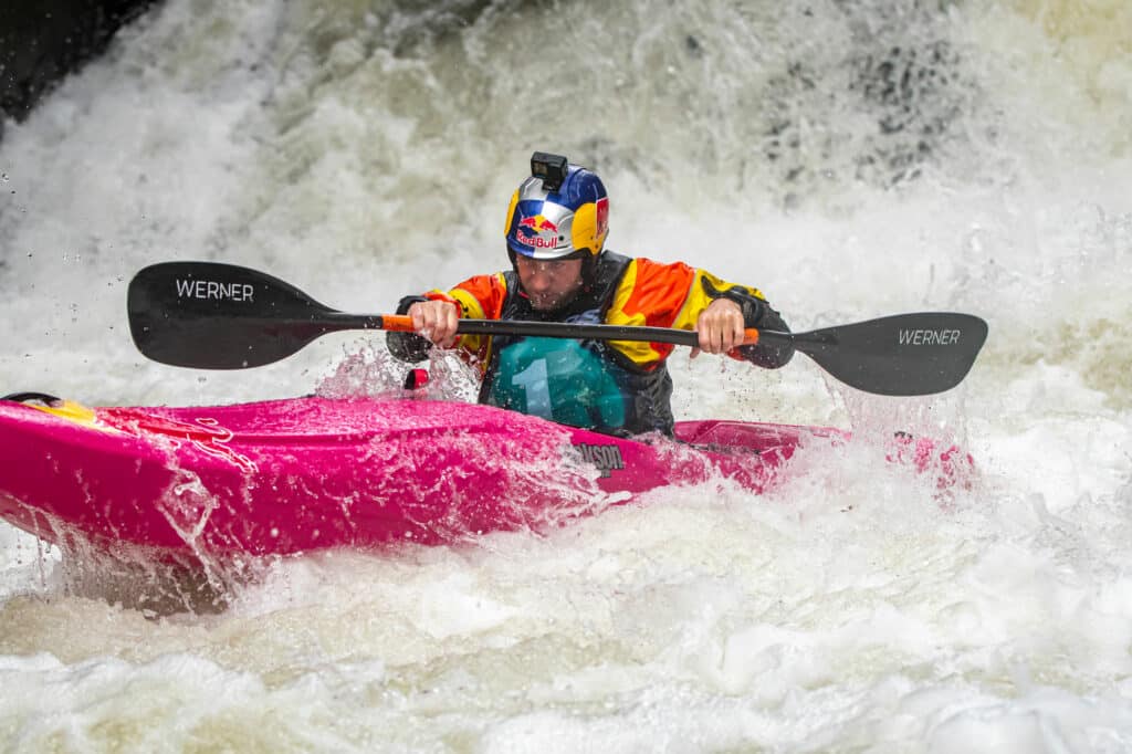 Dane Jackson paddles through huge whitewater rapid with a big drop now behind him while he is dripping in water.