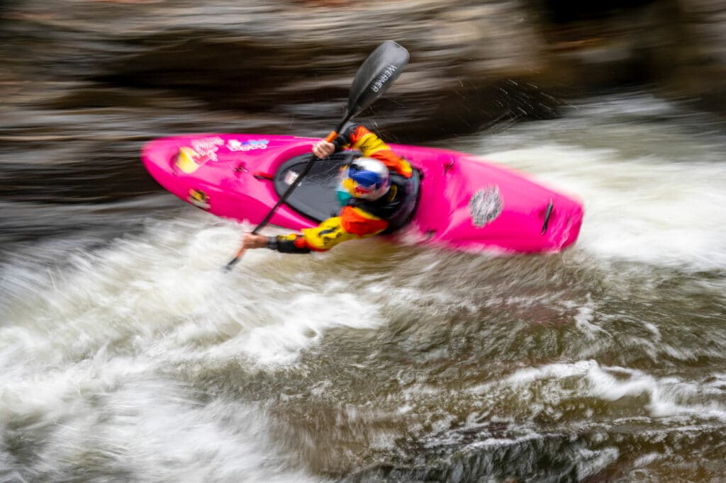 Blurred photo of Dane Jackson paddling his bright pink whitewater kayaking through fast moving and extreme whitewater rapids.
