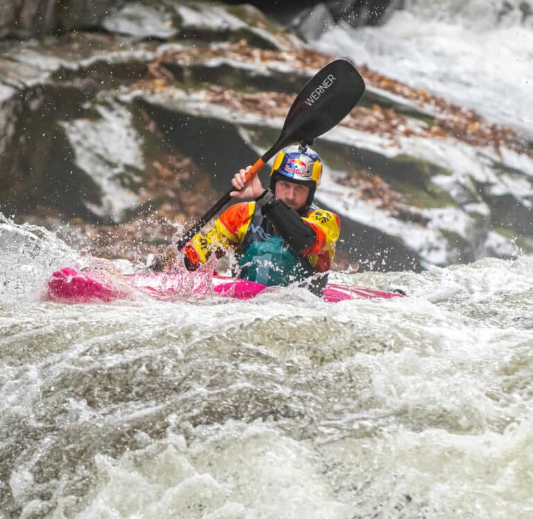 Dane Jackson starts to paddle into a big drop with huge whitewater rapids and rocks seen behind him.