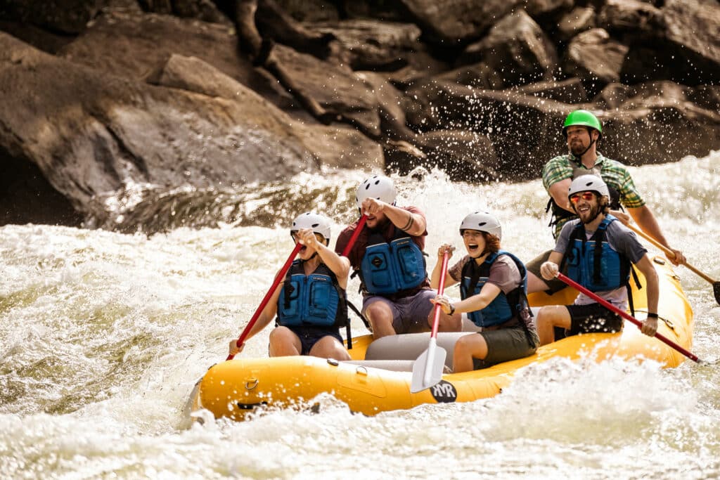 Five people in protective white water gear laugh and smile as they paddle through rushing white water in a yellow inflatable raft.