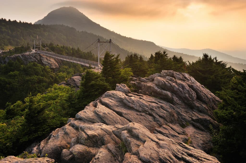A overlook view from Grandfather Mountain that shows lush forest, rolling mountains, and a white bridge during sunset.