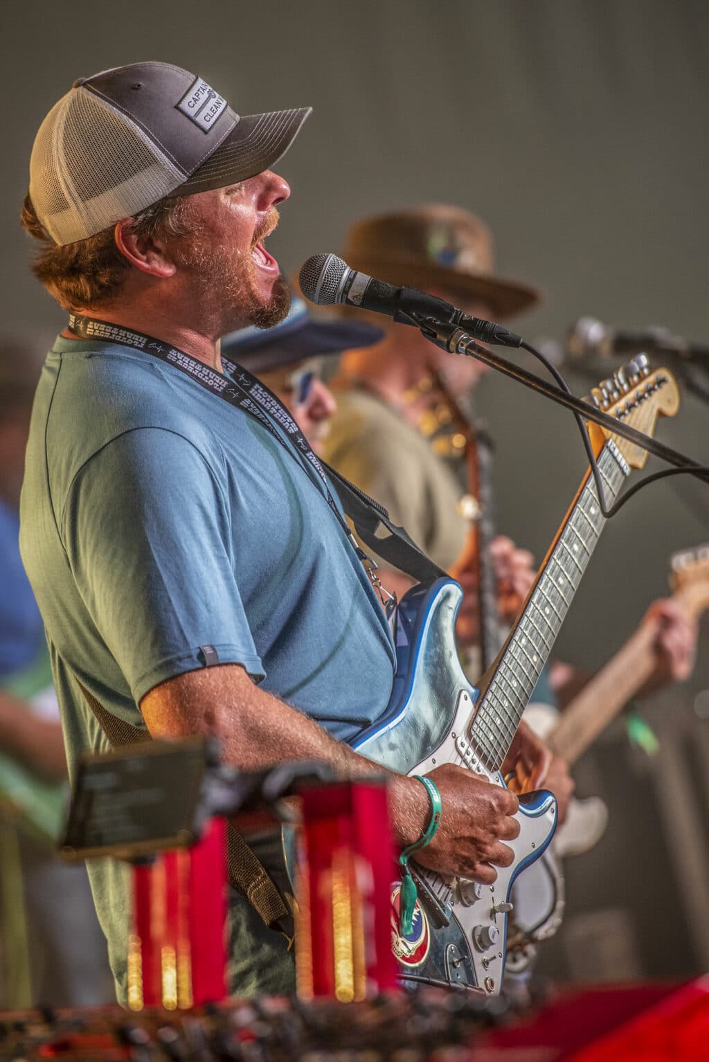 Bearded man playing an electric guitar sings into a microphone while wearing a truckers baseball cap.