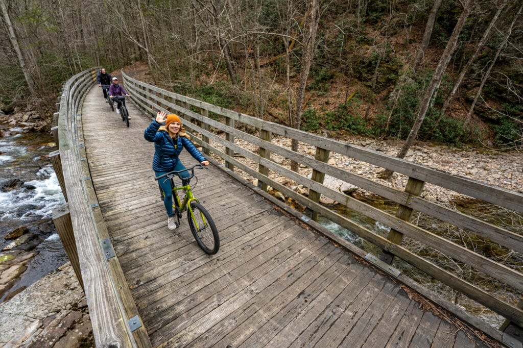 Women in blue jacket and orange hat waves as she rides her bike over a wooden bridge with two bikers behind her.