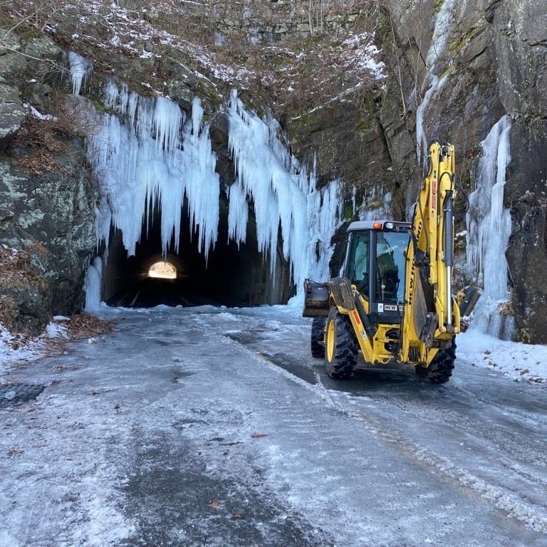 A yellow batco tractor is parked in front of a long road tunnel that is covered in thick icicles.