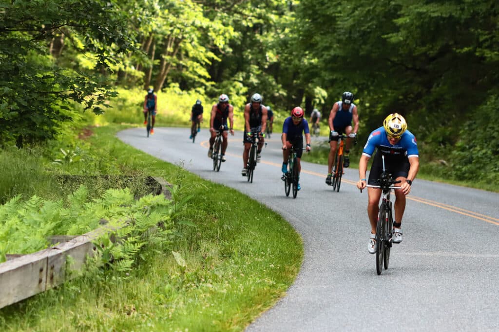 A group of triathletes in riding gear ride their road bikes on a paved road that winds through a green forest.