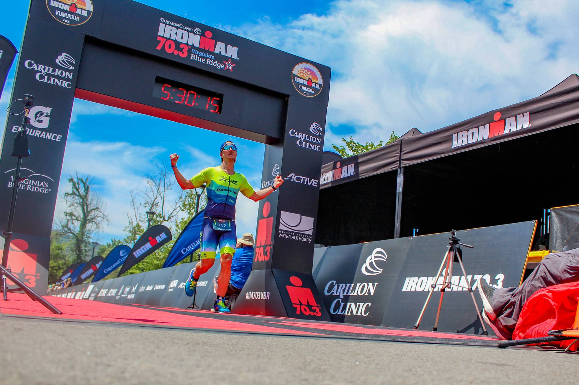 Marathon runner crosses the finish line with his arms up as he passes under the Ironman arch with a clock timing the race on display.