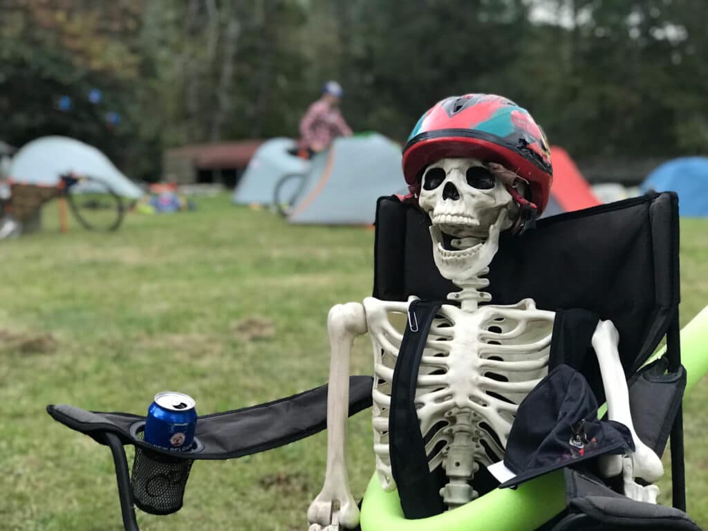 A decorative, plastic human skeleton sits in a camp chair at a campsite with tents while wearing a bike helmet and other recreational safety gear.