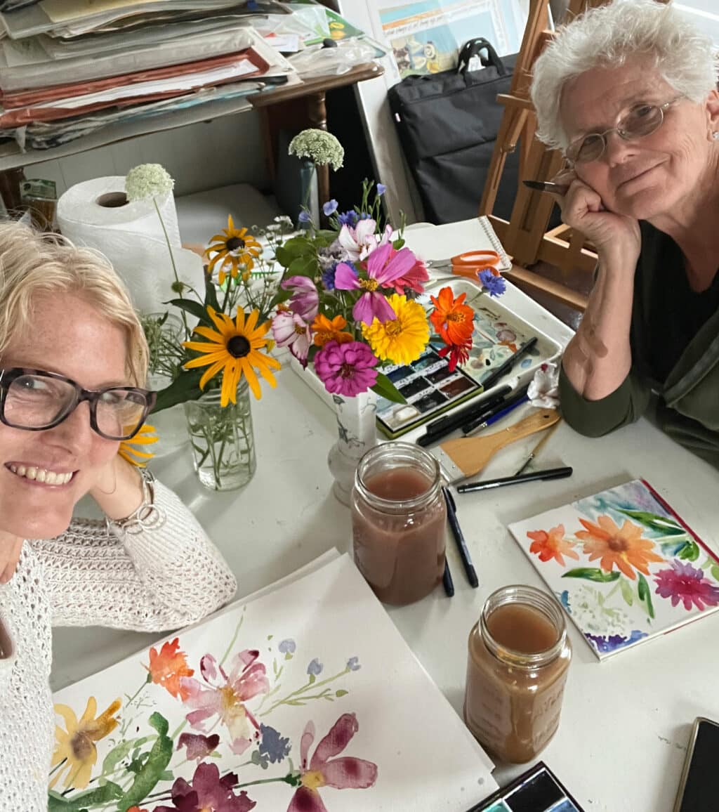 Holly, left, and Delia Wach paint wildflowers together at a table with colorful fresh picked flowers in a vase.