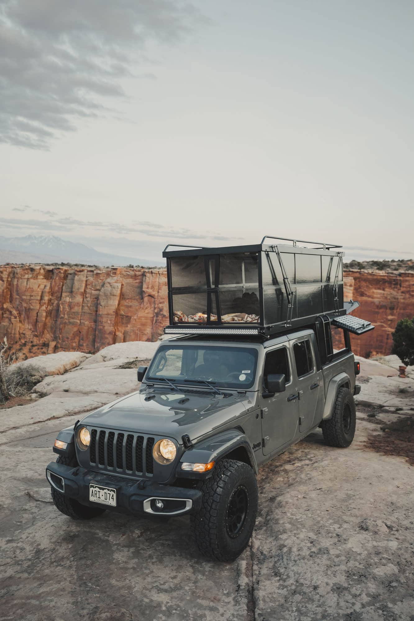 A jeep truck sits on top of a desert canyon with a camping pop-up attached to the roof.