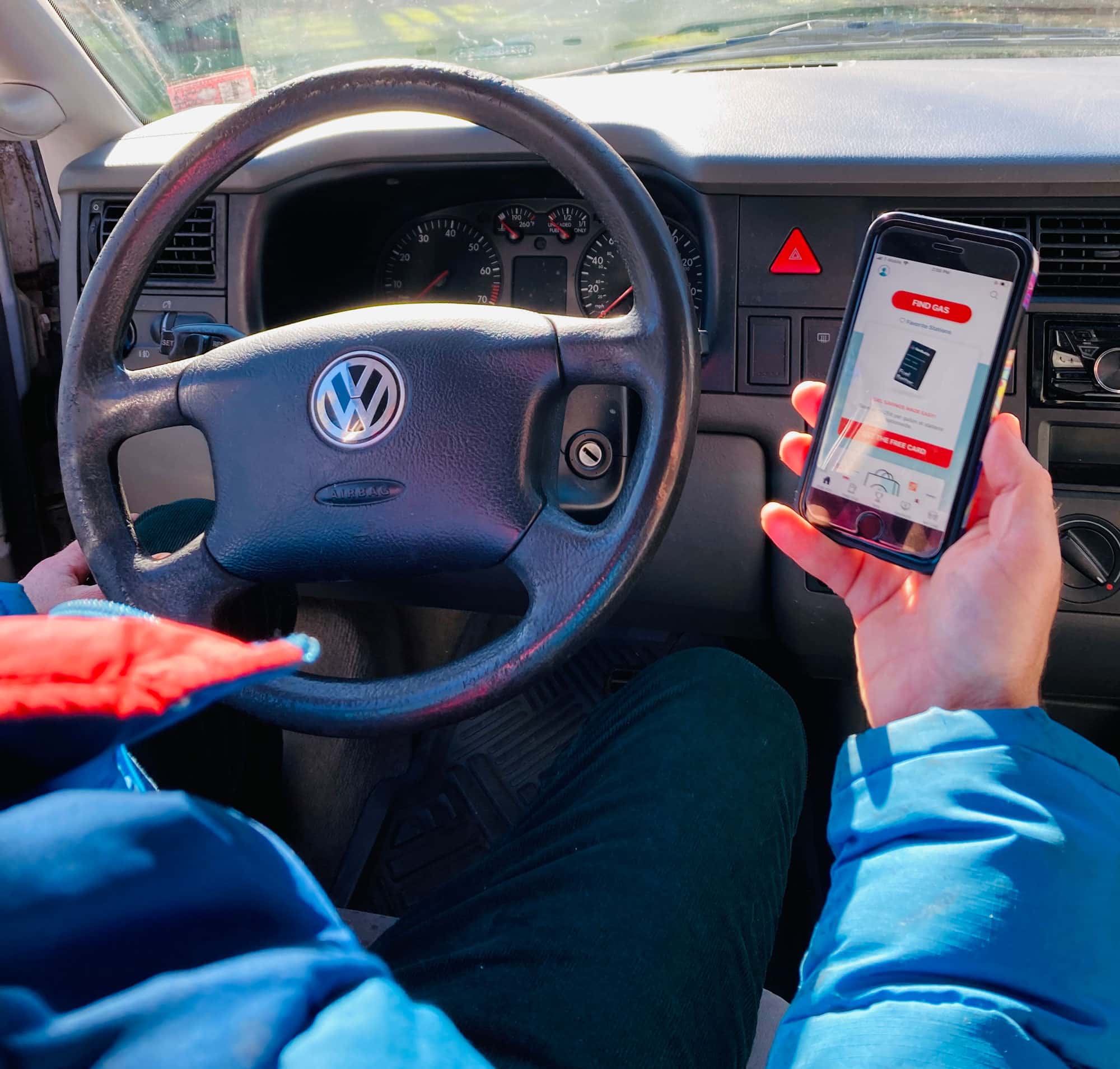 View of person in blue puffy jacket while they sit behind the wheel of a Volkswagon car and read something on their phone.