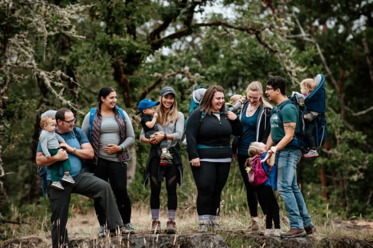 group of moms stand smiling while holding their children in hiking back carriers