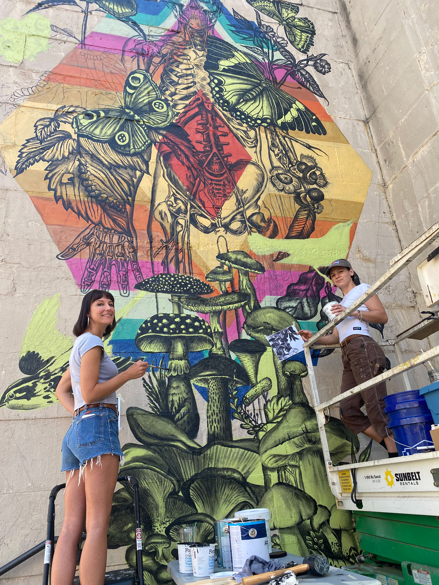 Two women stand in front of a colorful mural that they are painting. The mural contains bright colors of red, orange, pink, and yellow while depicting butterflies, moths, a human skeleton, and leaves.