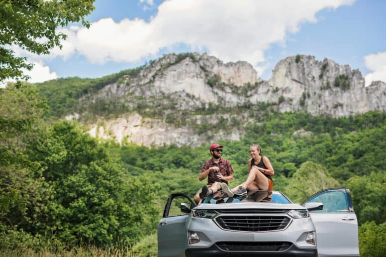two people sit on their car with the backdrop of a dramatic mountain range, Seneca Rocks.