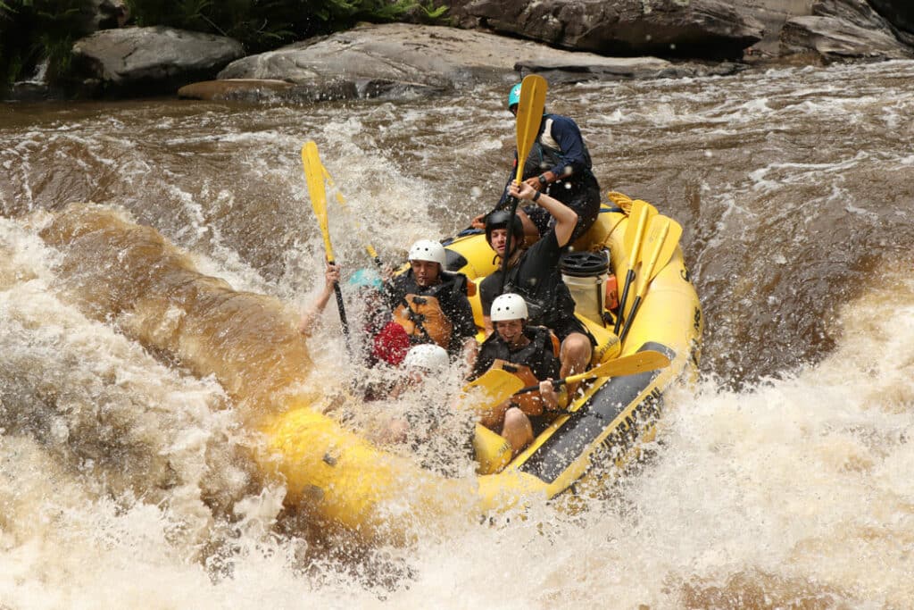 group of people wearing white water safety gear smile and yell as they go through big whitewater rapids in a yellow inflatable raft.