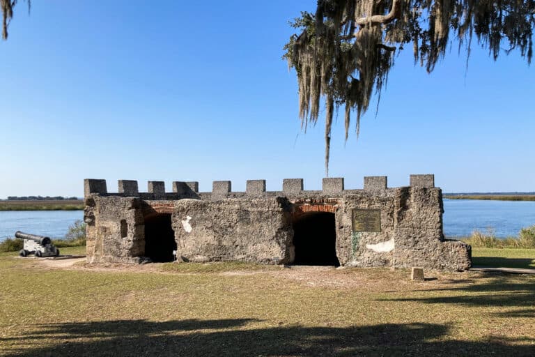 Stone castle-shaped fort stands under a Floridian tree next to a body of water in the sunshine.