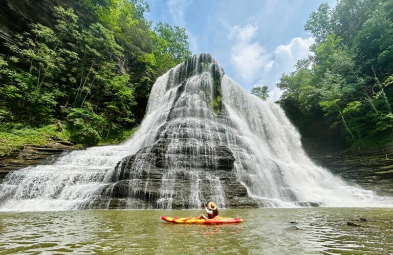 Person kayaking looks up at a big waterfall they are under in a forest