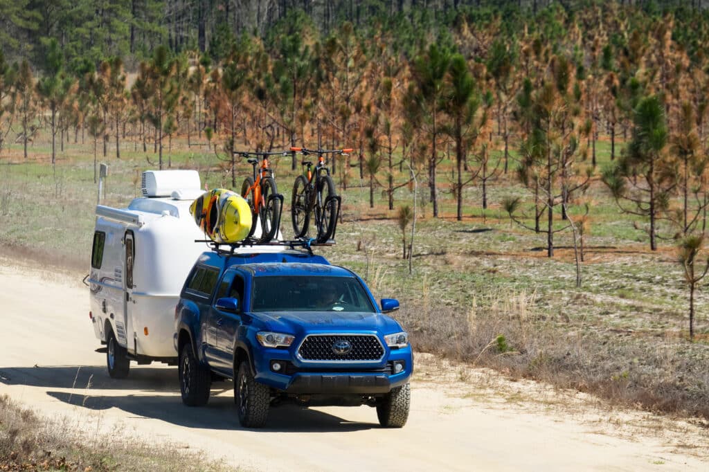 Blue Toyota truck carries two mountain bikes on top of it, a kayak behind it, and towing a small white camper.